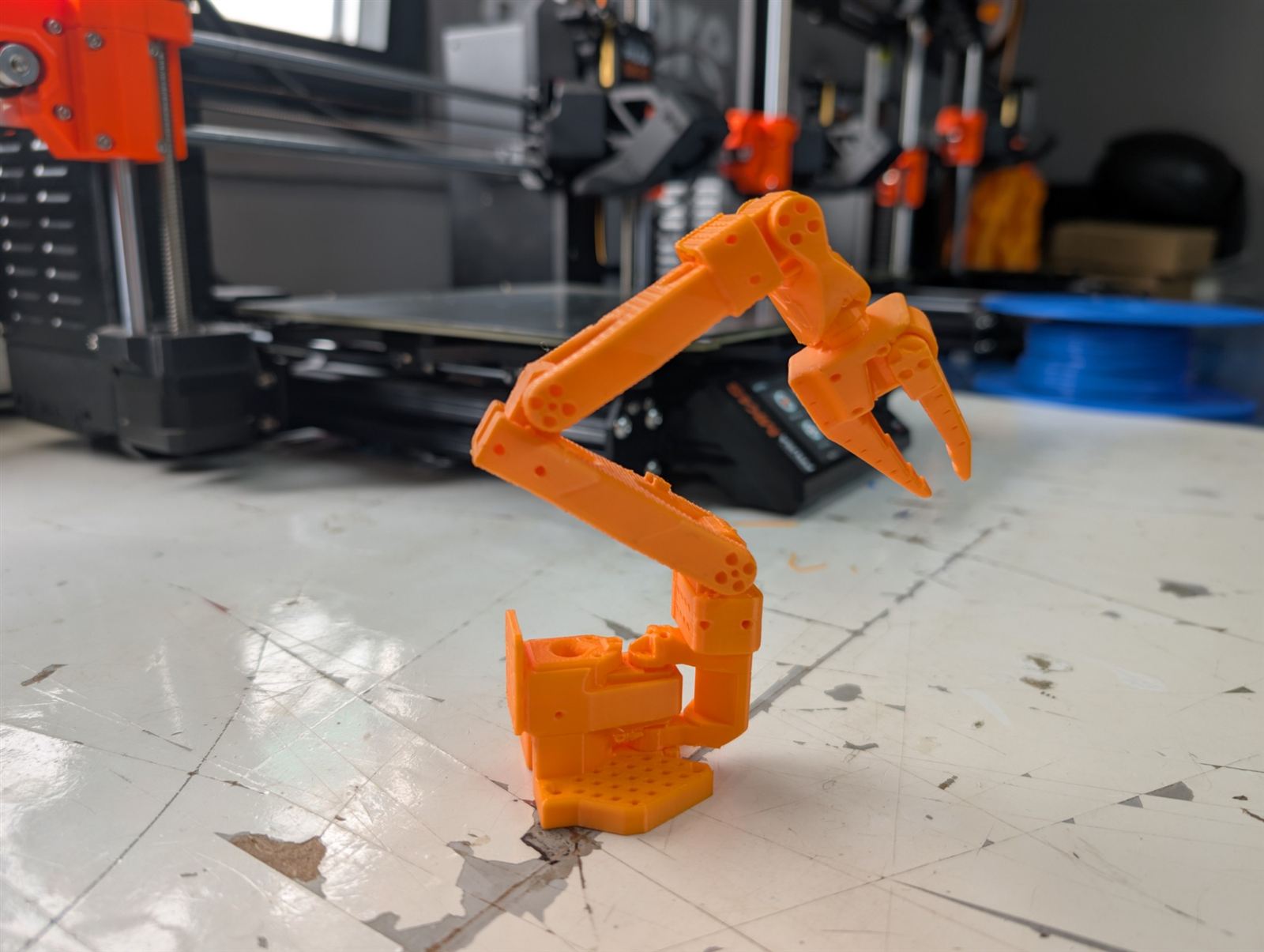 Un bras SO-ARM imprimé en 3D en PLA orange, devant les imprimantes de l'atelier